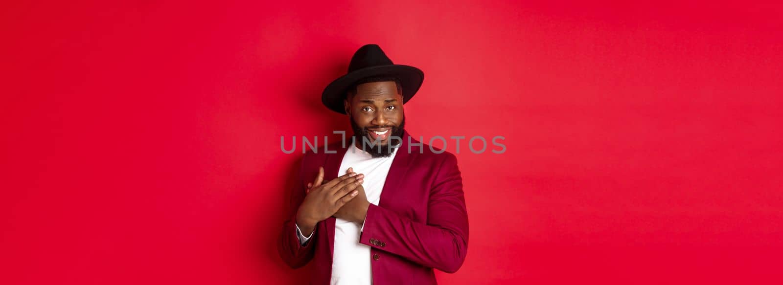 Fashion and party concept. Handsome african american macho man flirting, smiling and holding hands on heart, being grateful and touched, standing over red background.