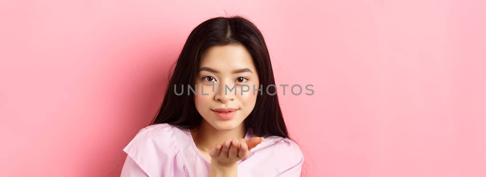 Beautiful asian girl with natural makeup, blowing air kiss at camera with sensual gaze, standing romantic against pink background by Benzoix