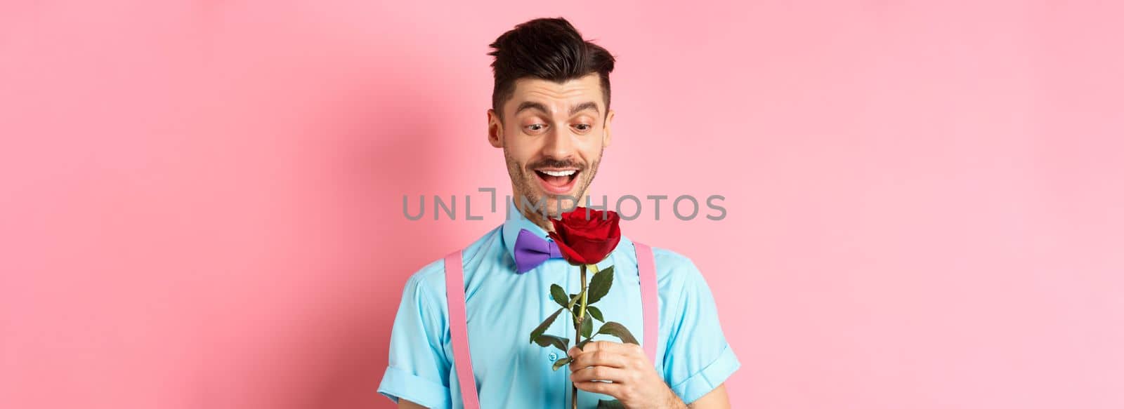 Valentines day and romance concept. Happy boyfriend looking at red rose while waiting for lover in bow-tie, standing on pink romantic background.
