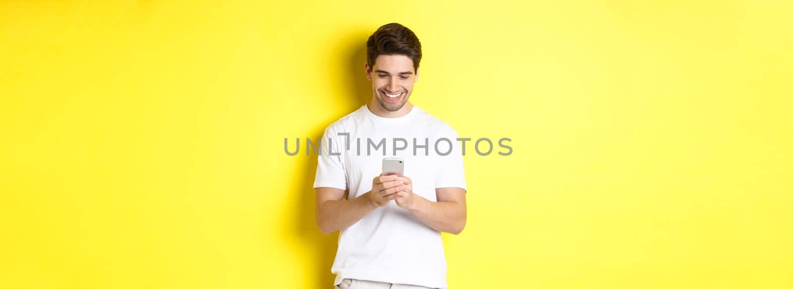 Young man reading text message on smartphone, looking at mobile phone screen and smiling, standing in white t-shirt against yellow background.