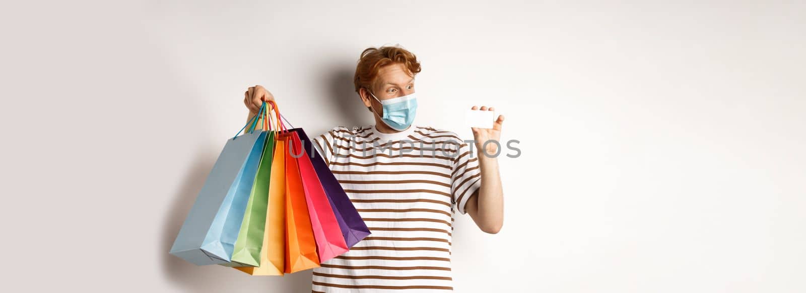 Pandemic and special offer concept. Happy redhead man in face mask showing shopping bags and plastic credit card, standing over white background.