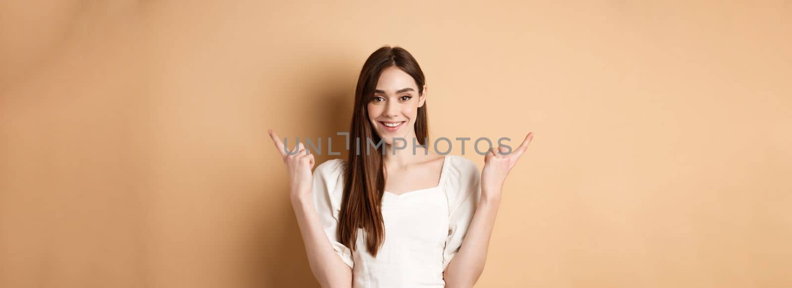 Beautiful young woman in dress pointing fingers sideways, show two ways, giving choice and smiling, standing on beige background by Benzoix