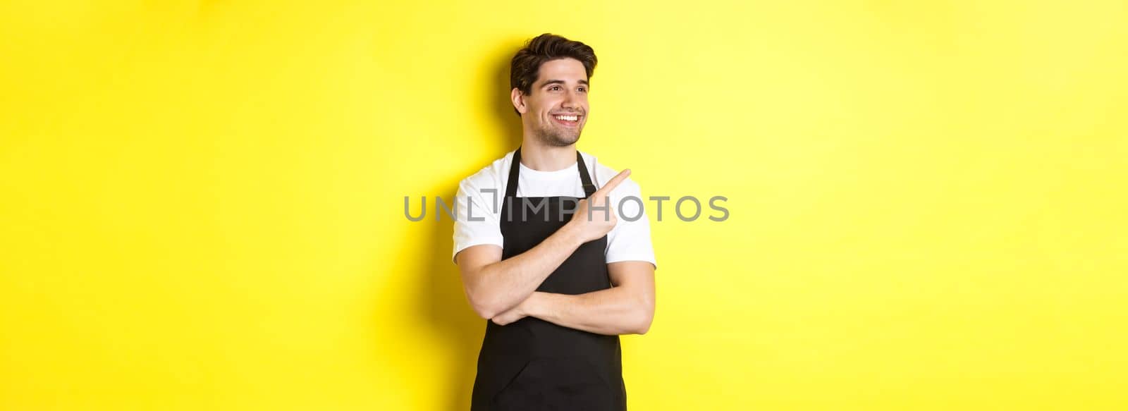 Handsome barista pointing and looking left at promo, wearing black apron, standing against yellow background.