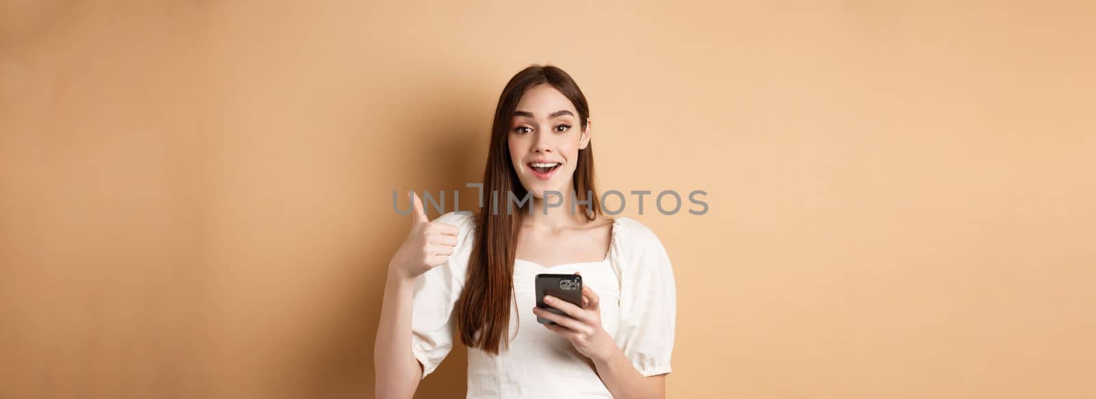 Online shopping concept. Happy young woman showing thumb up and using cellphone, smiling pleased, standing on beige background by Benzoix