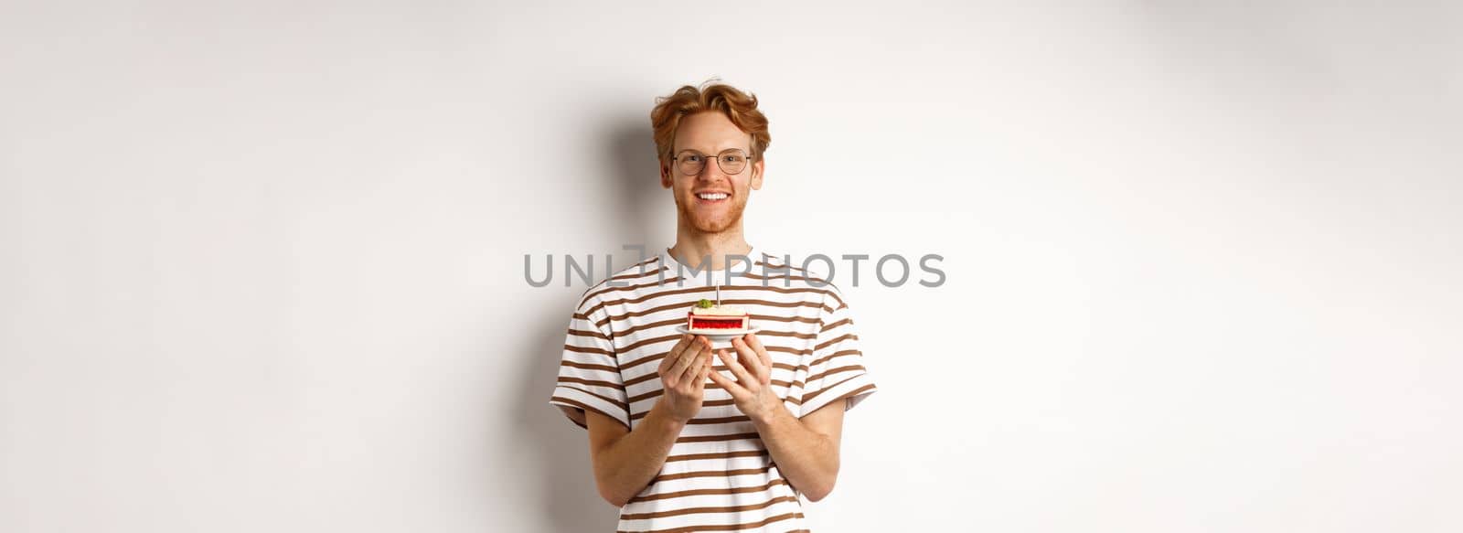 Holidays and celebration concept. Handsome redhead man in glasses holding birthday cake with candle, smiling and looking happy at camera by Benzoix