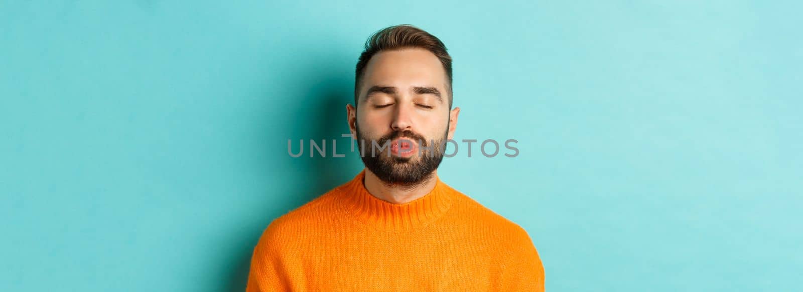 Close-up of young man in sweater pucker lips, close eyes and waiting for kiss, standing over light blue background.