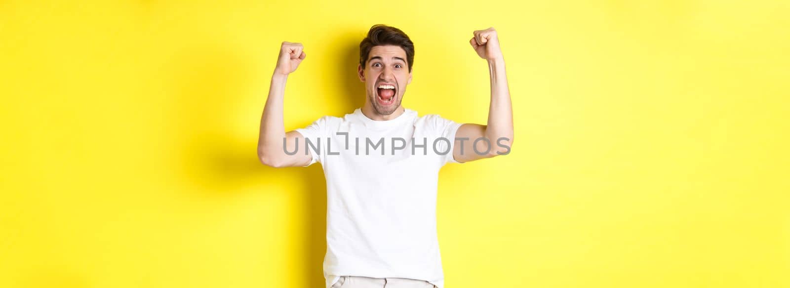 Image of excited man winning, raising hands up and celebrating, triumphing and rooting for team, standing over yellow background by Benzoix