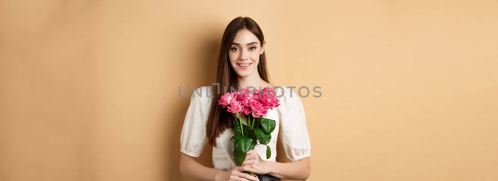 Romantic Valentines day concept. Beautiful young lady holding pink roses and smiling, standing happy on beige background.