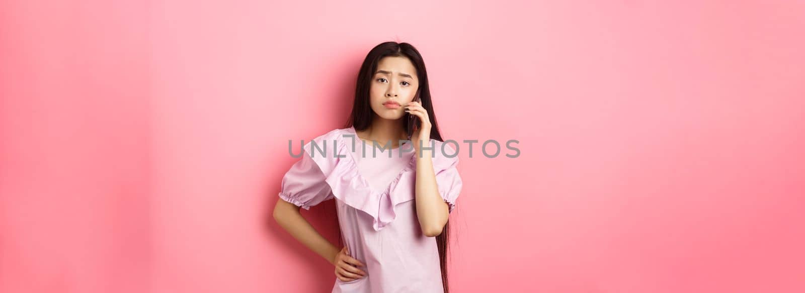 Sad asian girl calling someone, holding phone and talking, standing upset against pink background by Benzoix