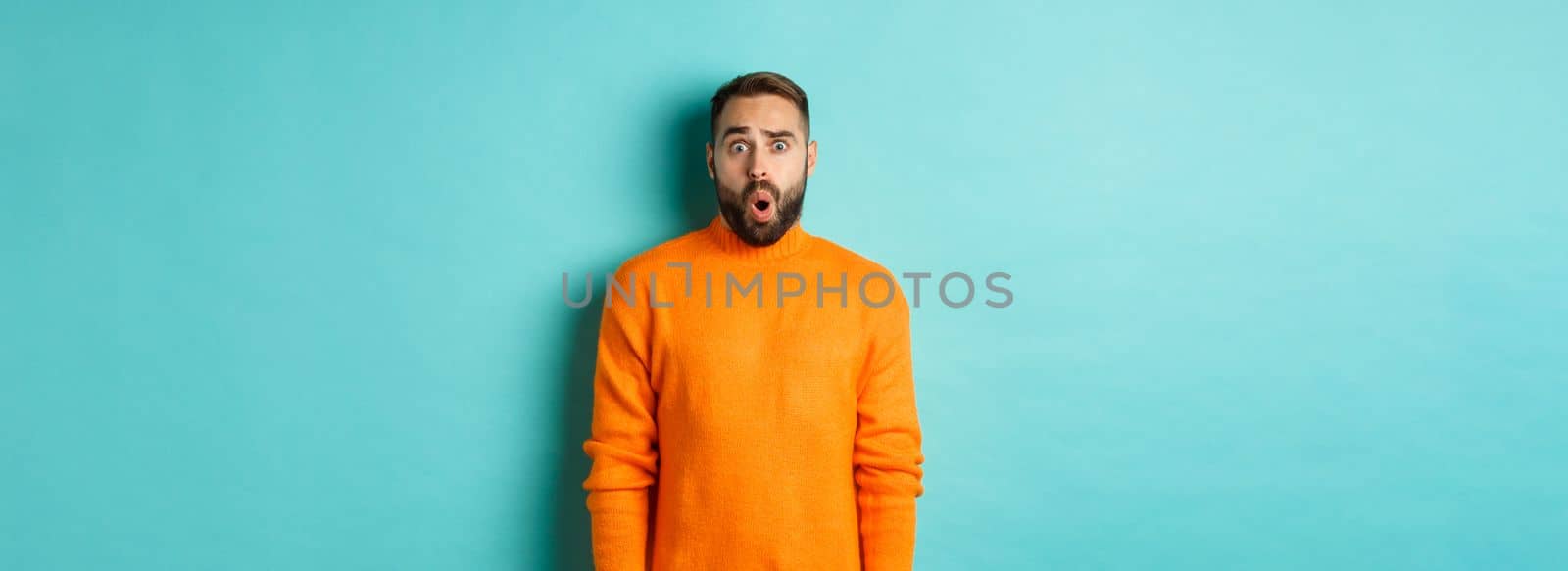 Shocked and confused man looking in awe at promo offer, standing near copy space on turquoise background.