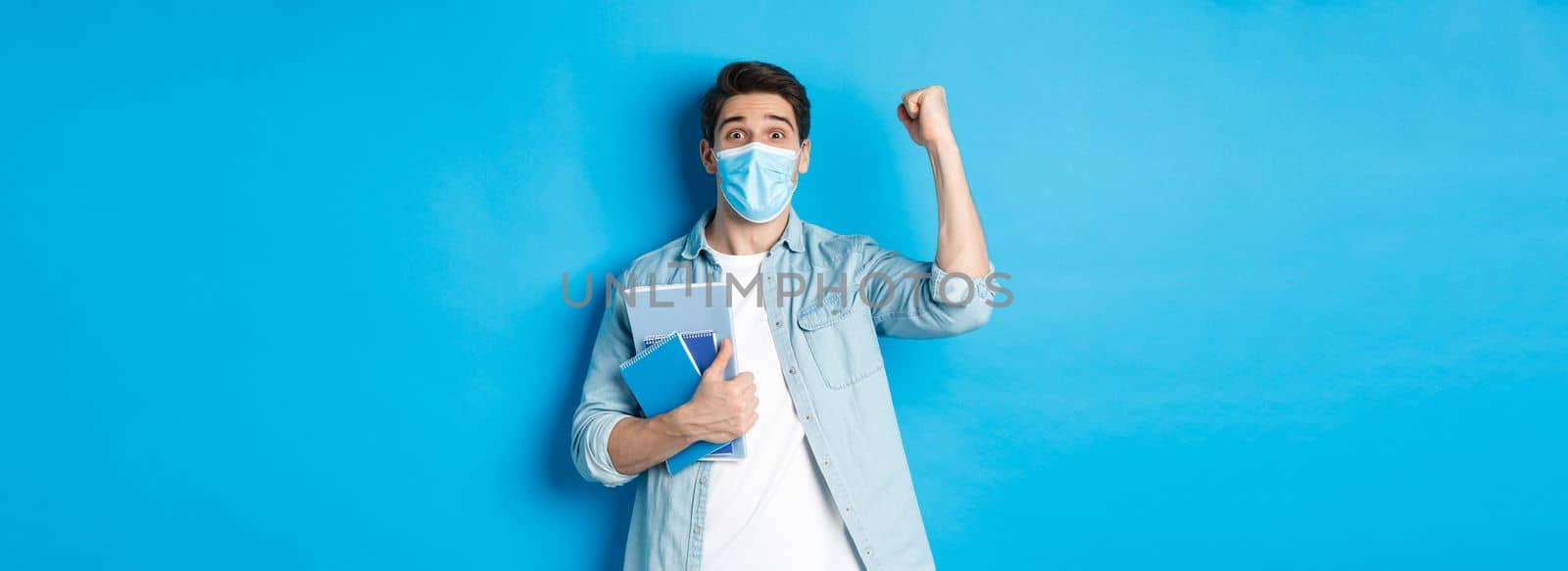 Education, covid-19 and social distancing. Excited male student in medical mask triumphing, raising fist up and holding notebooks, standing over blue background.