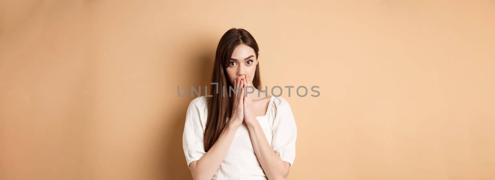 Omg shocking. Speechless woman covering mouth with hands and staring shocked at camera, gossiping, hear stunning news, beige background by Benzoix