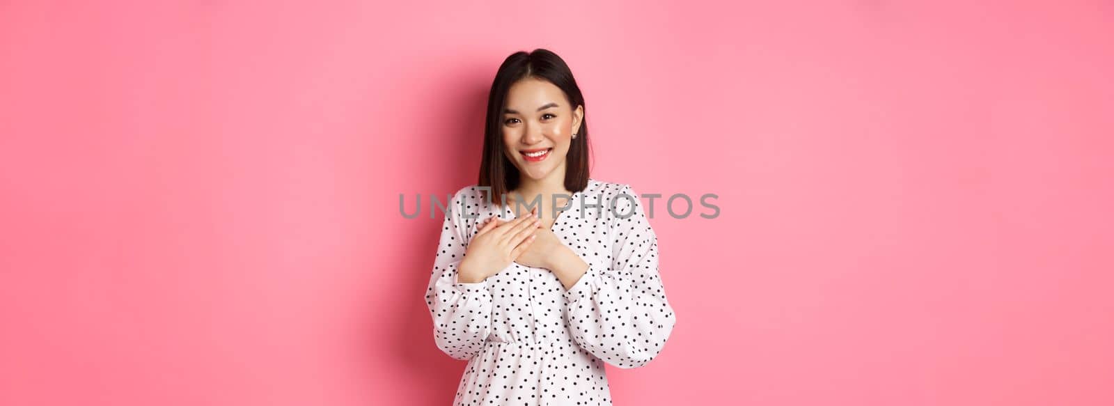 Thankful Korean girl in dress smiling, holding hands on heart and looking grateful at camera, touched with nice gesture, standing over pink background by Benzoix