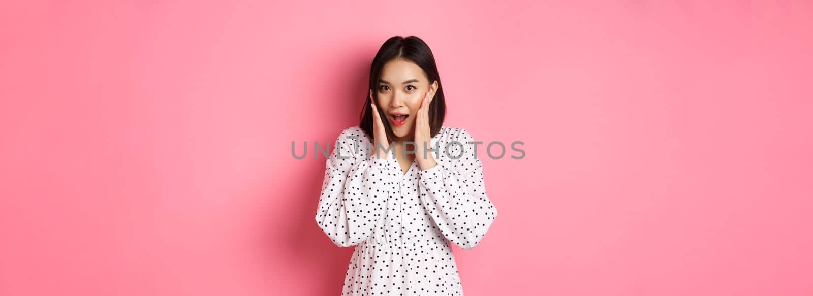 Young asian woman looking intrigued, gossiping and saying wow with curious face, looking interested at camera, standing over pink background by Benzoix