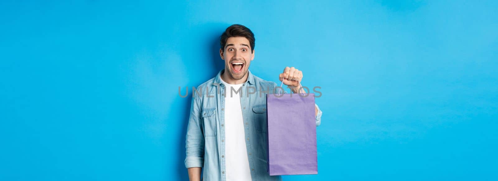 Concept of shopping, holidays and lifestyle. Excited handsome guy holding paper bag with gift and looking happy, standing over blue background.