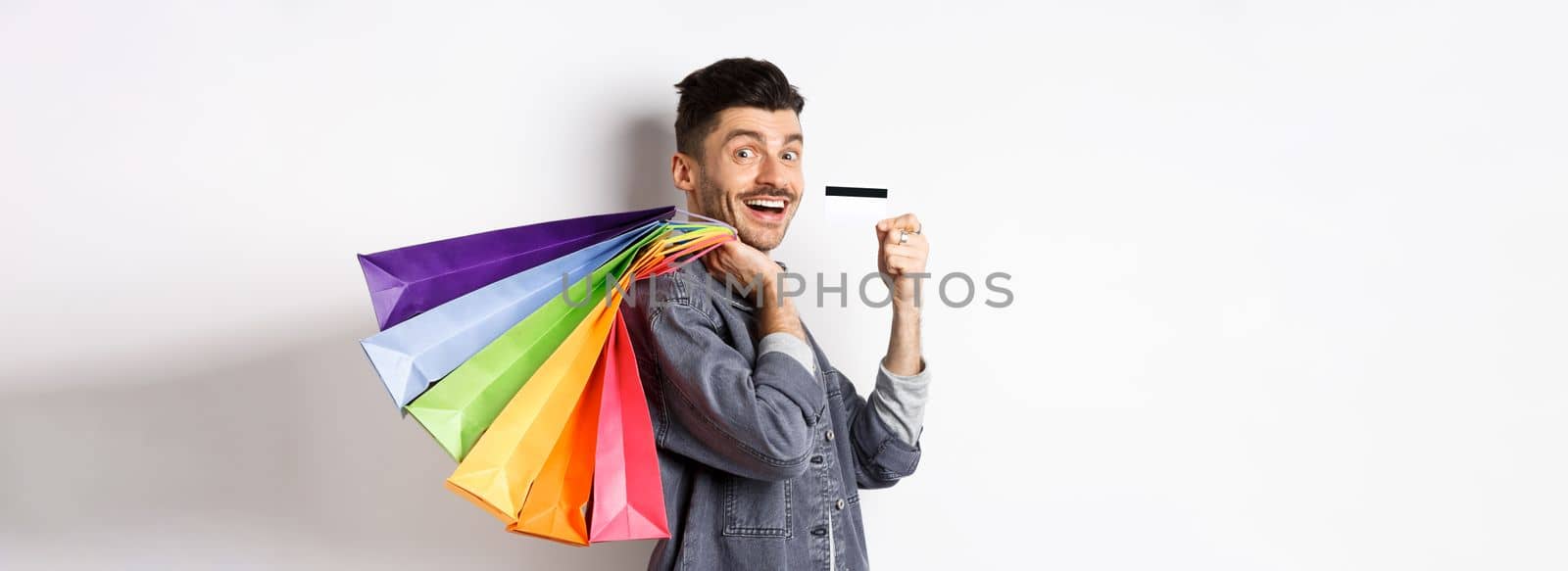 Cheerful guy going on shopping with credit card, holding bags on shoulder and smiling excited at camera, white background.
