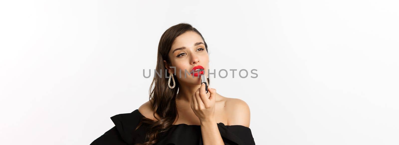 Fashion and beauty concept. Beautiful woman in black dress applying red lipstick and makeup, going on party, standing over white background.