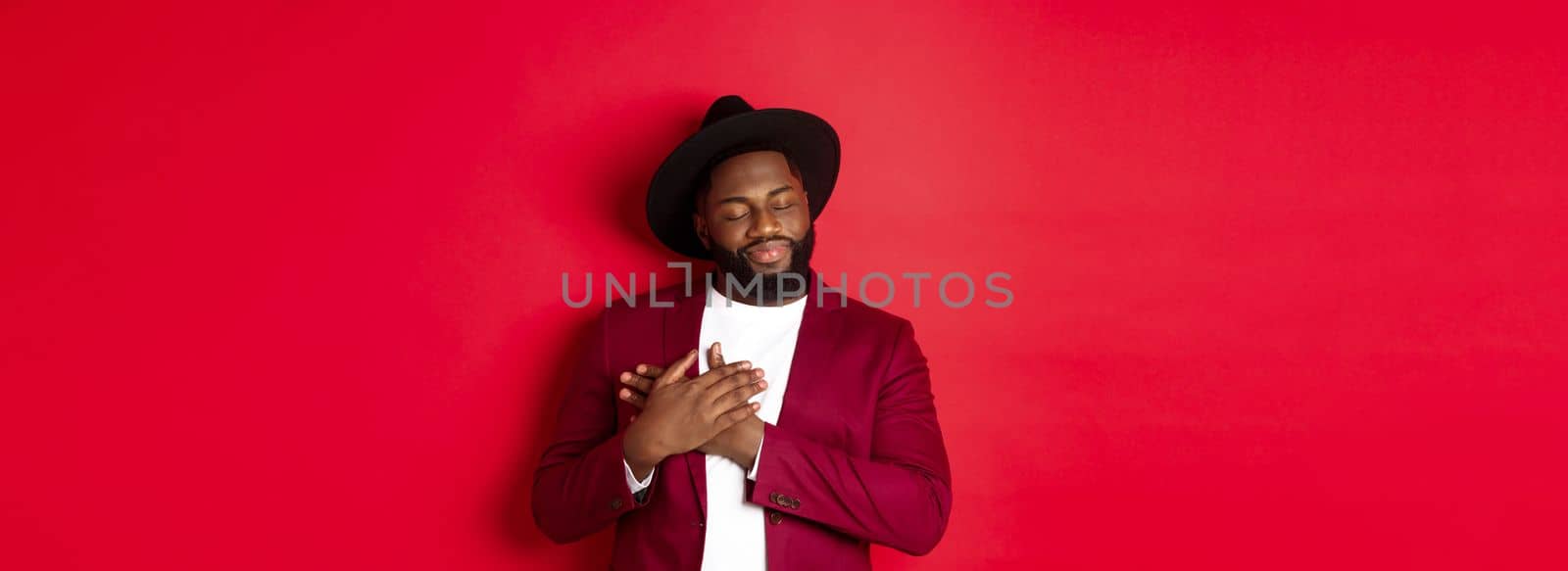 Fashion and party concept. Young bearded Black man in classy hat and jacket, holding hands on heart and smiling nostalgic, remember something, daydreaming against red background.