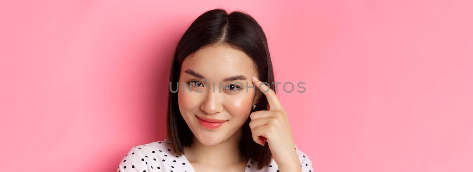 Beauty and skin care concept. Headshot of smart asian woman pointing at head and smiling sly, asking to think, standing over pink background.