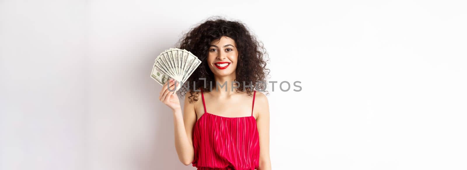 Stylish smiling woman in red dress showing money, standing on white background by Benzoix