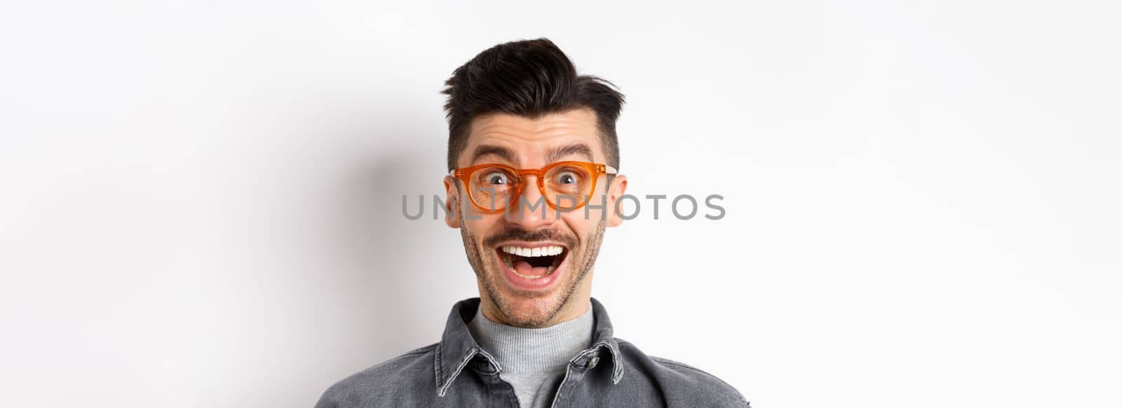 Excited funny man in glasses smiling, look with amazement and joy at awesome promo, standing happy against white background.