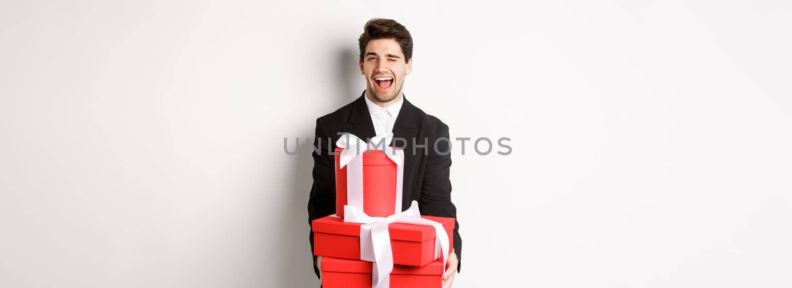 Image of handsome guy in black suit, holding gifts and winking at camera, have christmas presents, standing against white background by Benzoix