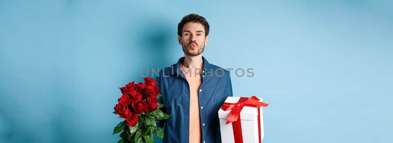 Love and Valentines day concept. Romantic boyfriend pucker lips for kiss, bring bouquet of red roses and gifts on date, standing over blue background.