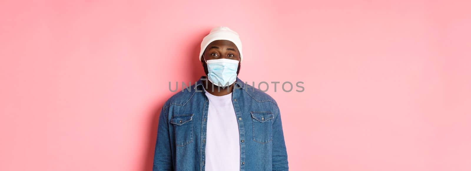 Covid-19, lifestyle and quarantine concept. Happy Black guy in beanie and face mask smiling with eyes, standing over pink background.