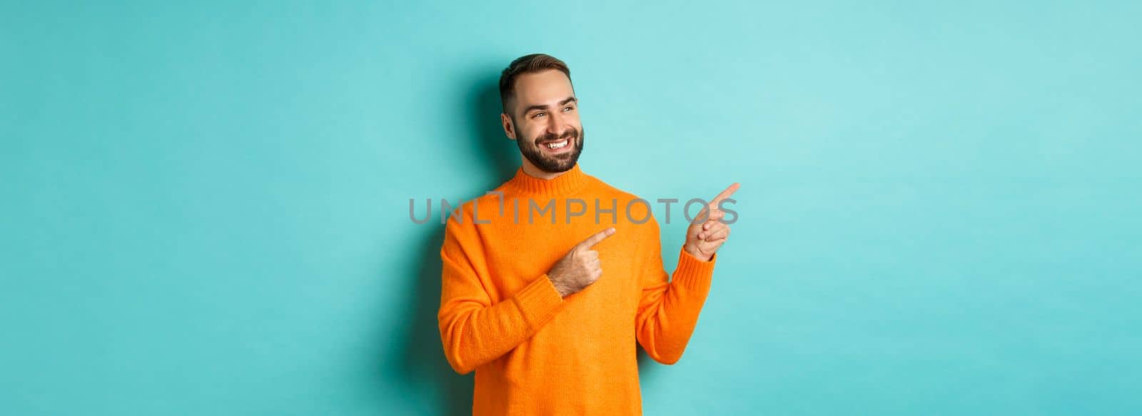 Attractive bearded male model in orange sweater, smiling satisfied and pointing fingers left, standing over turquoise background by Benzoix