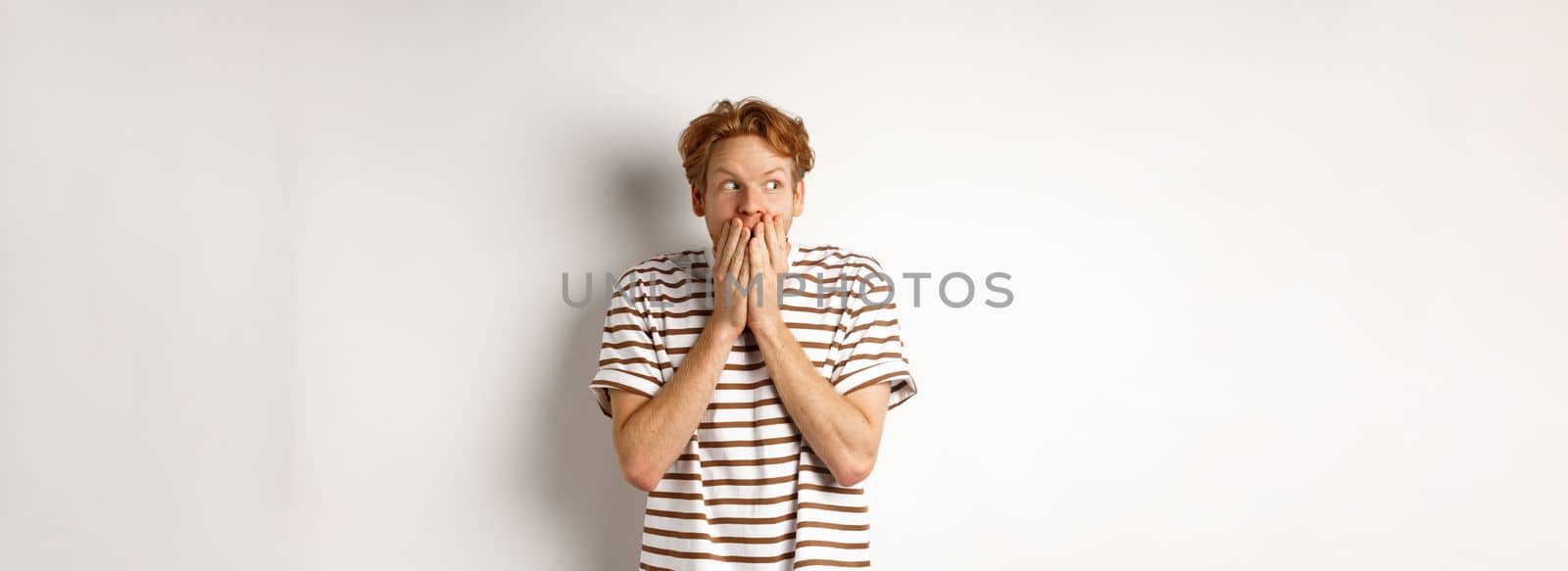Shocked redhead guy gossiping, giggle in hands and looking left impressed, standing over white background.
