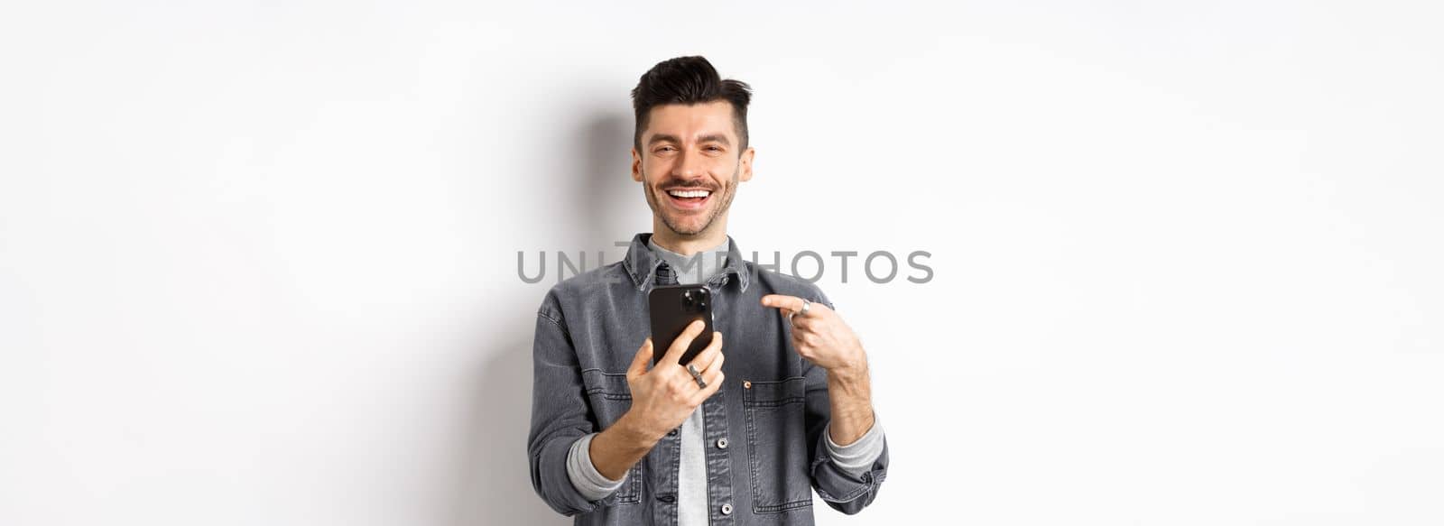 Happy handsome caucasian man with moustache pointing finger at smartphone, smiling and showing good deal online on mobile phone, standing pleased against white background.