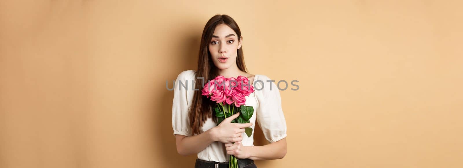 Valentines day. Image of surprised girlfriend thanking for flowers, receive pink roses from lover and looking grateful at lover, standing on beige background.