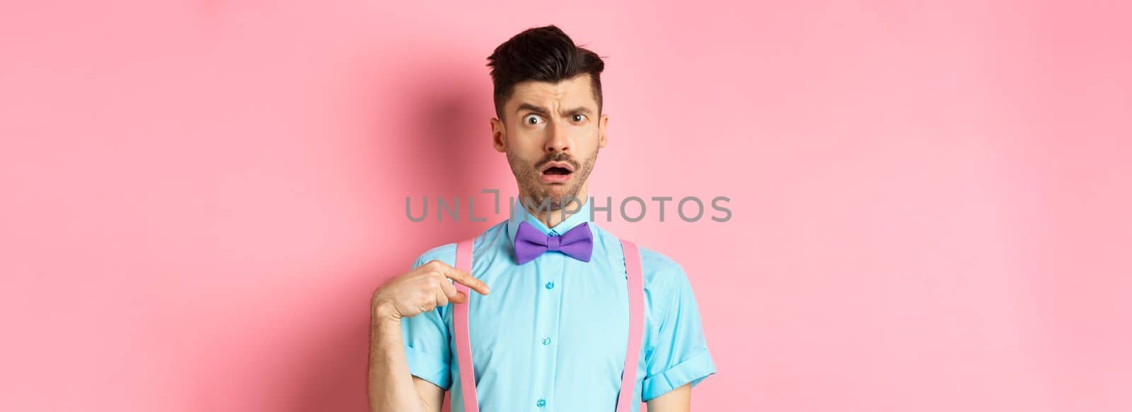 Image of confused funny guy in bow-tie and suspenders, pointing at himself as if being accused or chosen, raising eyebrow surprised, standing over pink background.
