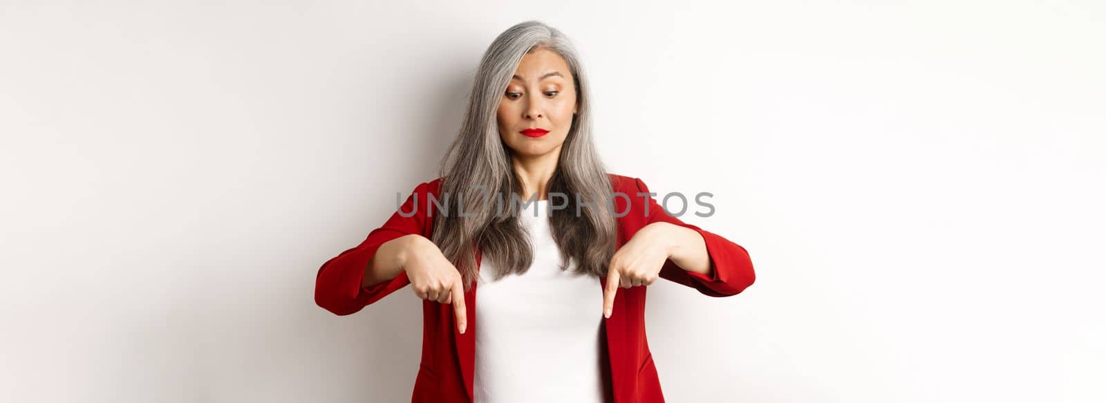 Intrigued asian elegant woman looking and pointing fingers down, showing logo, standing in red blazer over white background.