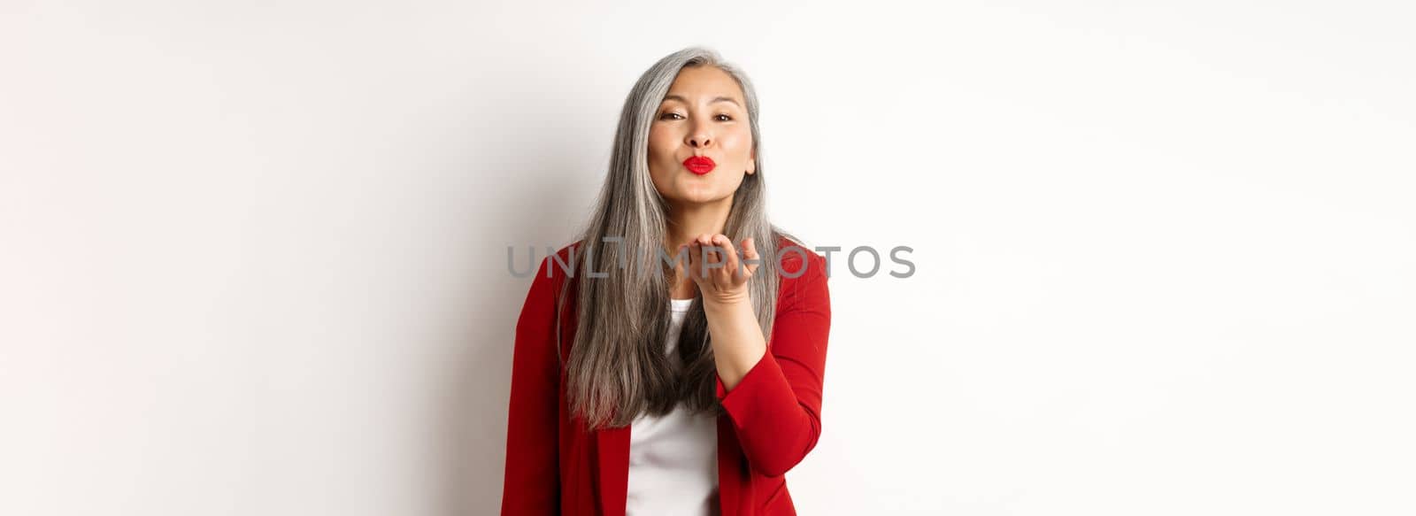 Elegant asian woman with red blazer and lips, blowing air kiss at camera, concept of valentines day and romance, white background.