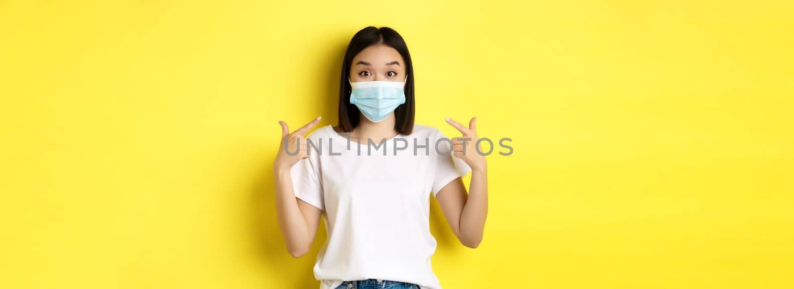 Covid-19, quarantine and social distancing concept. Young asian woman in white t-shirt, pointing at her medical mask from coronavirus, yellow background.