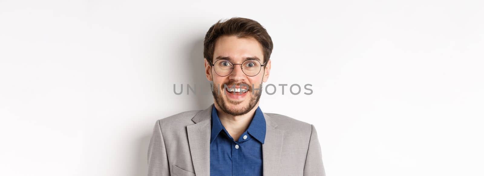 Cheerful european man in glasses and suit smiling excited, looking at camera with amazed happy face, white background by Benzoix