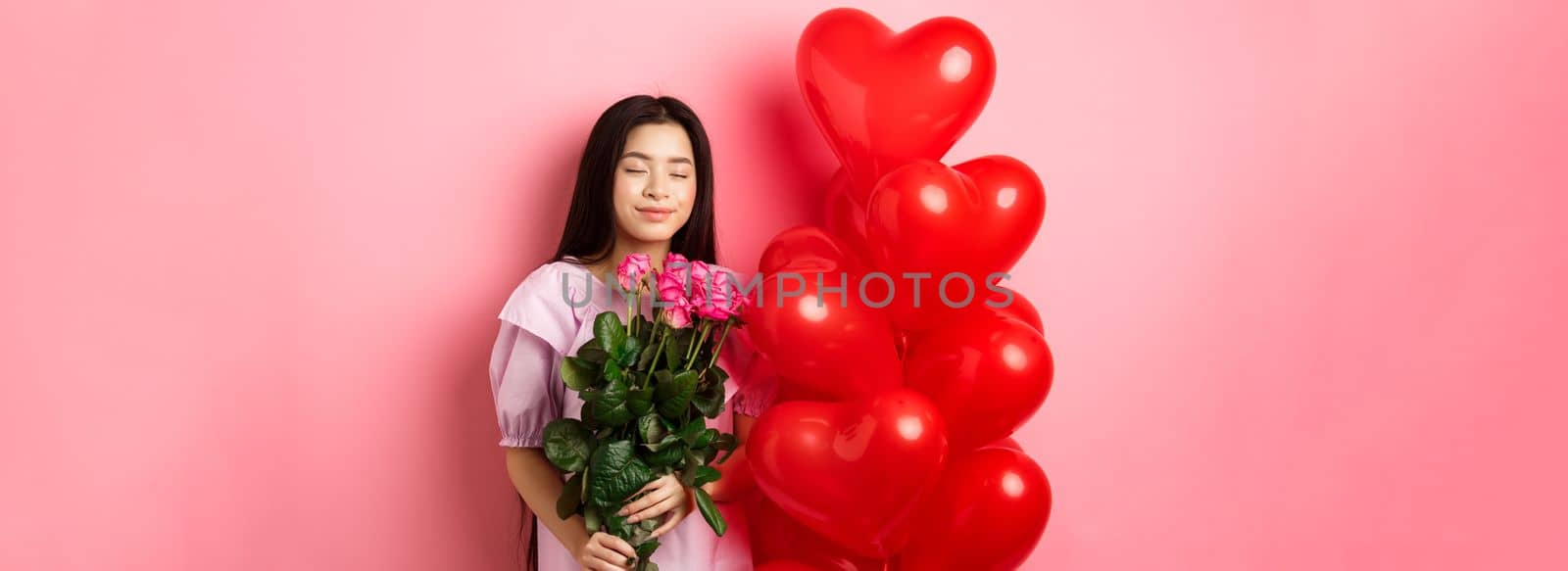 Valentines day concept. Romantic teen asian girl dreaming of love or date, close eyes and smile, holding flowers from lover, receive bouquet of roses and red heart balloons, pink background by Benzoix