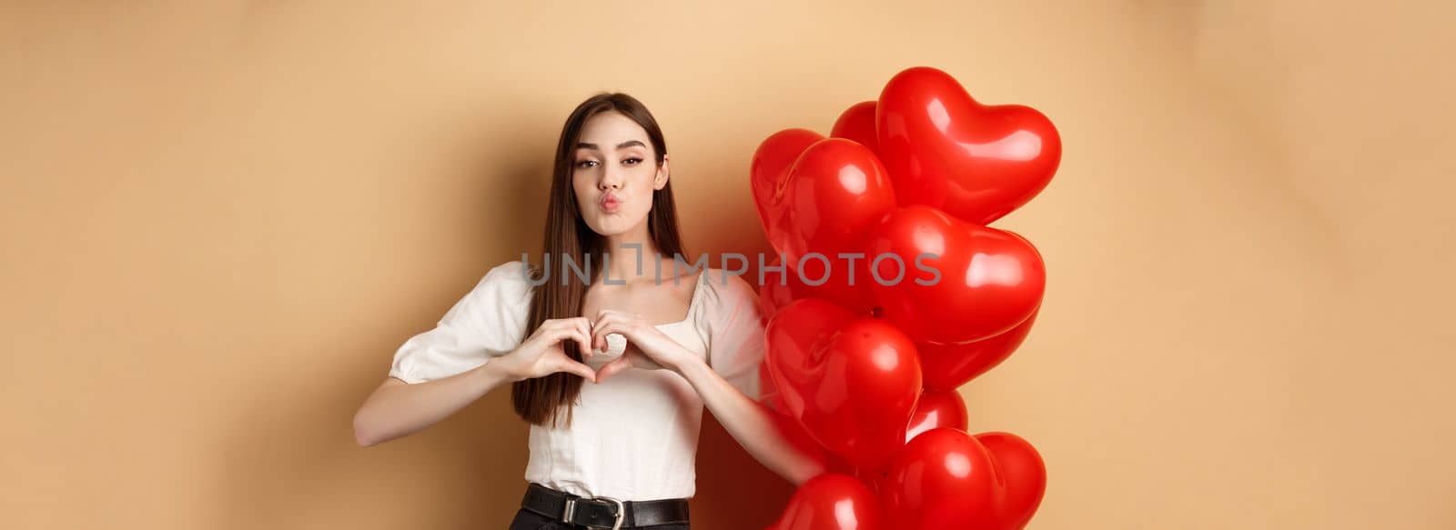 Lovely girlfriend pucker lips and waiting for romantic kiss, showing heart gesture to say I love you, look at lover, standing near Valentines day balloons and beige background by Benzoix