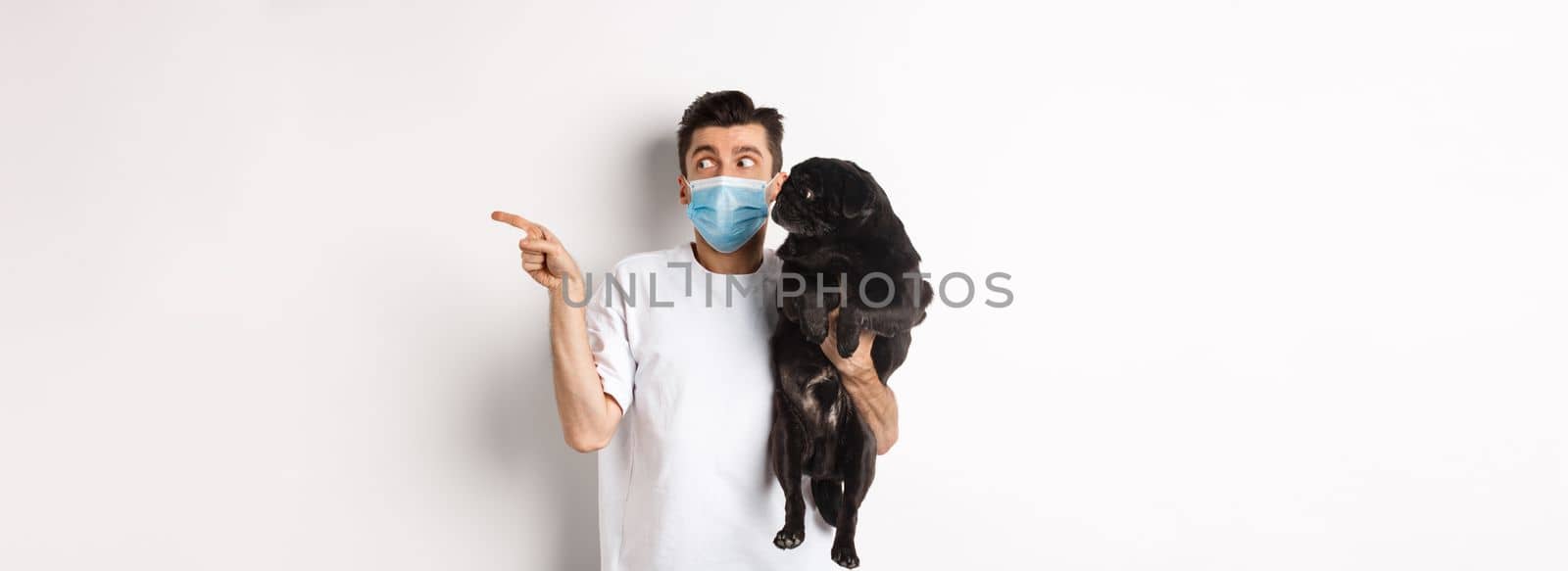 Covid-19, animals and quarantine concept. Young man in face mask holding cute black pug, dog and pet owner looking right at copy space, standing over white background by Benzoix