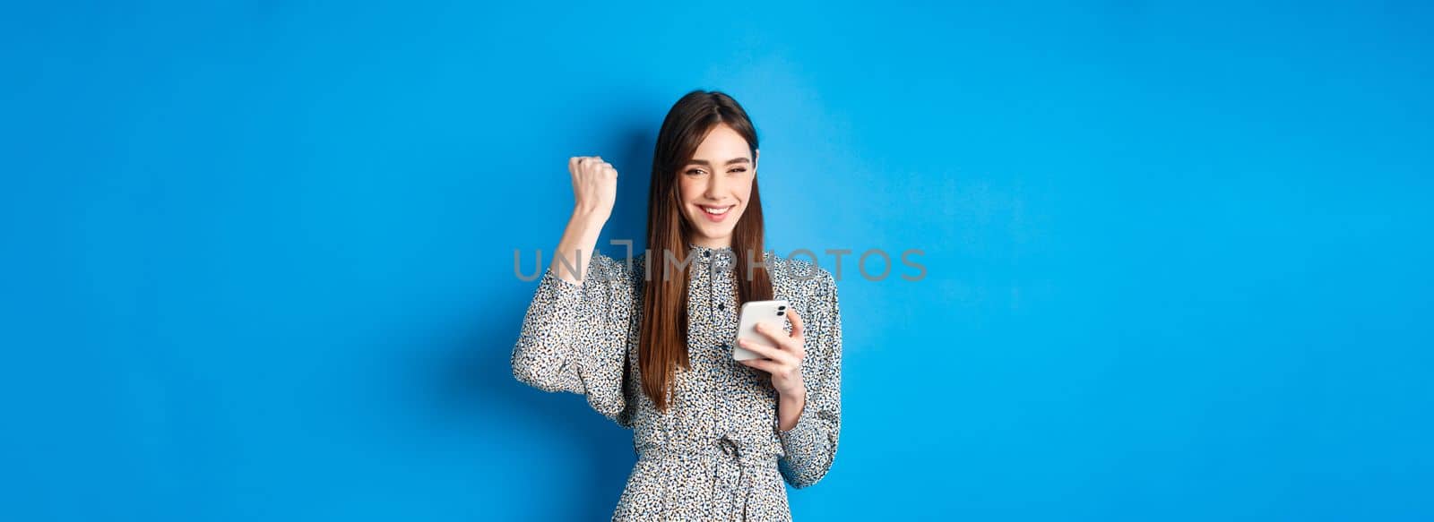 Happy pretty girl winning on mobile phone, smiling and chanting, holding smartphone, standing in dress against blue background.
