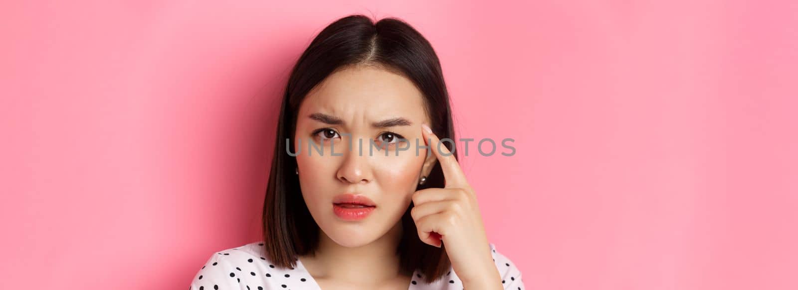 Beauty and skin care concept. Headshot of cute asian woman staring confused, pointing finger at head at frowning, standing over pink background by Benzoix