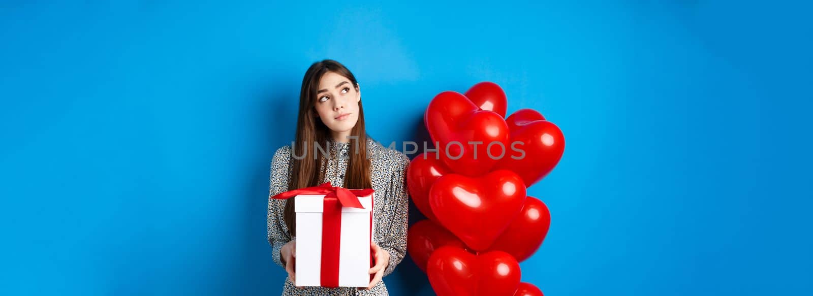 Valentines holiday concept. Pensive young woman in dress, holding gift and looking at upper left corner, thinking about lovers day, standing near red hearts balloons, blue background.