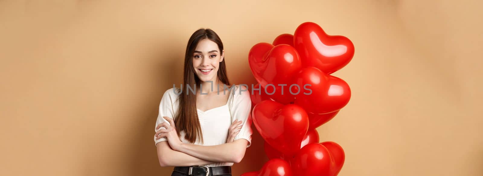 Cheerful young woman looking happy on Valentines day, standing near hearts balloons with arms crossed, smiling at camera, standing on beige background.