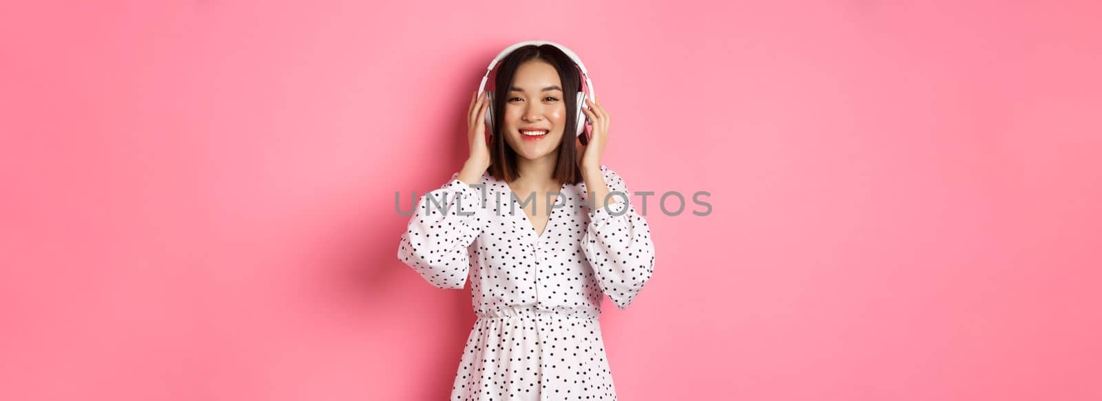 Romantic asian woman smiling happy, listening music in headphones and looking at camera, standing over pink background.