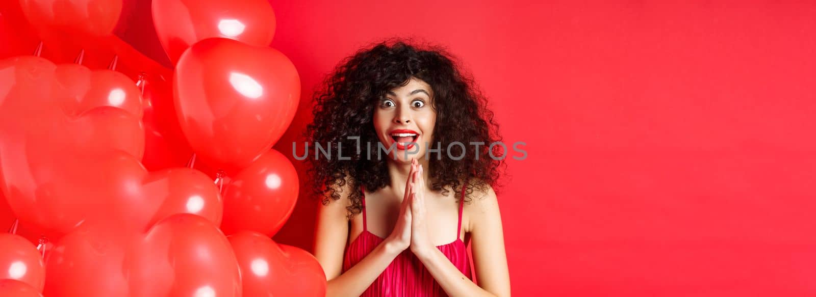 Valentines day. Excited caucasian woman in dress jumping from amazement, looking at something taunting, want to get product, standing near hearts balloons, white background.