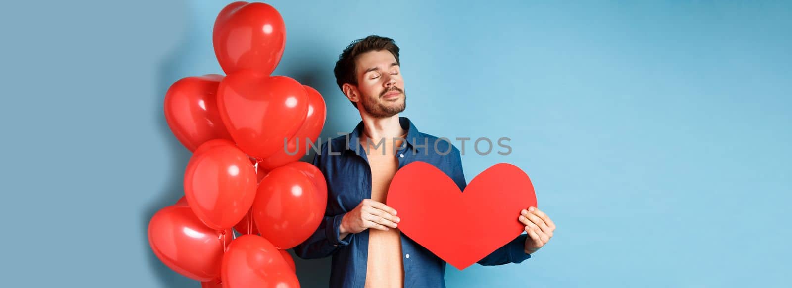 Valentines day and love concept. Dreamy man with closed eyes, holding romantic red heart cutout and standing near hearts balloons, blue background.