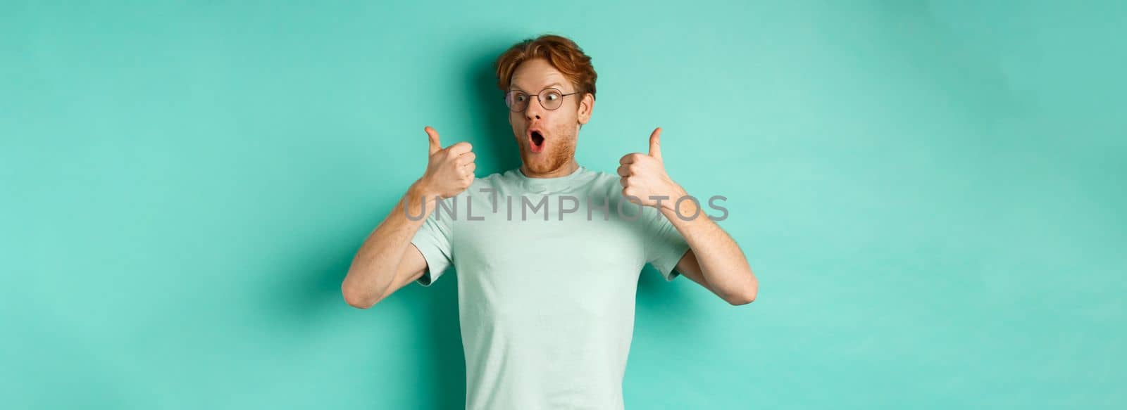 Excited young man with red hair and beard, staring at promo with awe, showing thumbs-up in approval, praising offer, standing over turquoise background by Benzoix