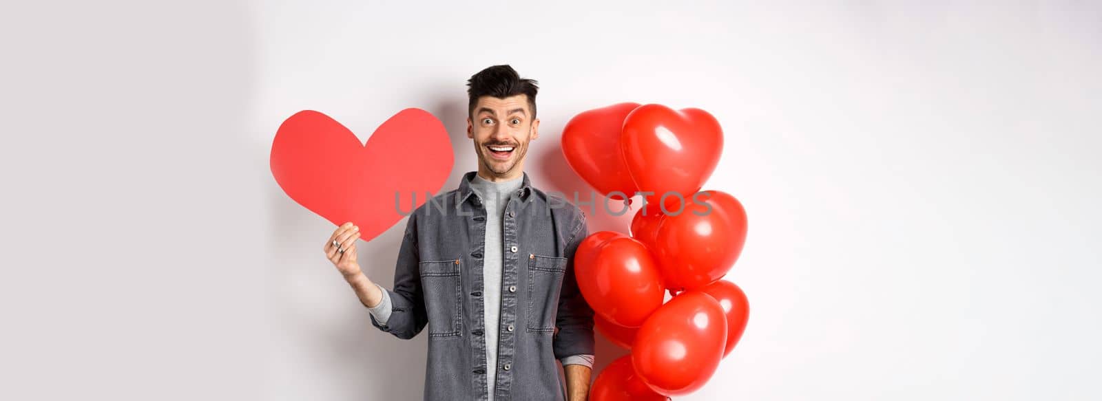 Valentines day and love concept. Cheerful funny guy showing heart cutout, standing near romantic balloons and smiling excited at camera, white background.