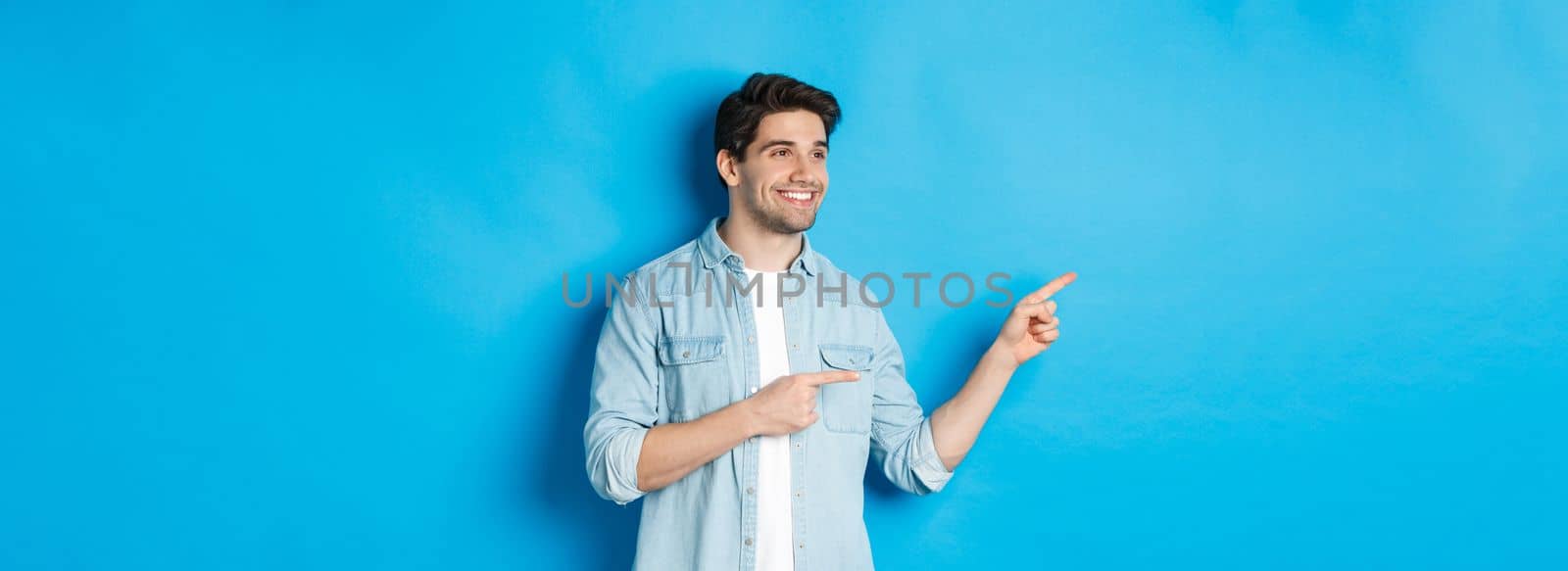 Handsome adult man introduce product, looking and pointing fingers left, promoting something against blue background.